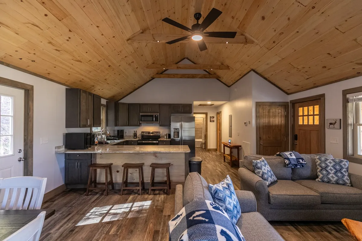 A wide view of the kitchen that flows into a cozy main living area, featuring a kitchen island, comfortable sofas, a wooden ceiling with a fan, and soft, natural light. The design highlights how the kitchen is the heart of the space, perfect for staying connected during meal prep and gatherings.