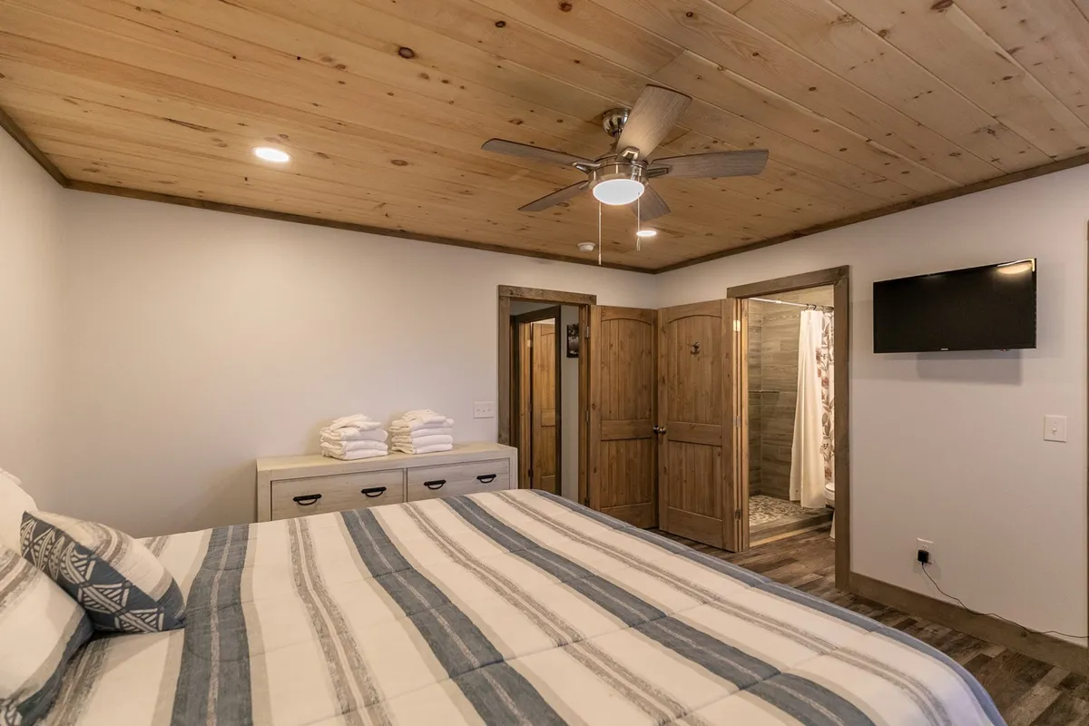 This image captures a cozy bedroom with a comfortable bed, wooden ceilings, a dressing area, and a television. The space feels warm and inviting, with natural textures highlighting the mountain-cabin ambiance while the modern amenities are subtly visible.