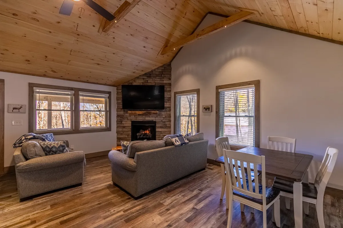 A spacious, open-concept living and dining area anchored by a stunning stone gas fireplace. Plush sofas and a dining table sit beside expansive windows that frame sweeping mountain views. The tongue-and-groove ceilings add rustic warmth, while natural light pours in, creating an inviting atmosphere for gatherings.