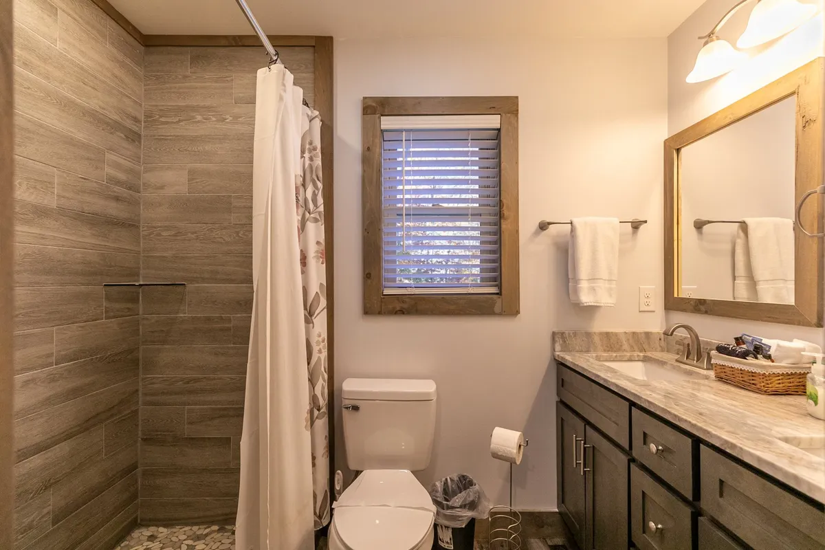 A bright and airy bathroom is shown, featuring a contemporary shower area, a sleek vanity with a sink, and a large mirror. The window with blinds allows natural light to filter in, giving the space a fresh, spa-like feel.