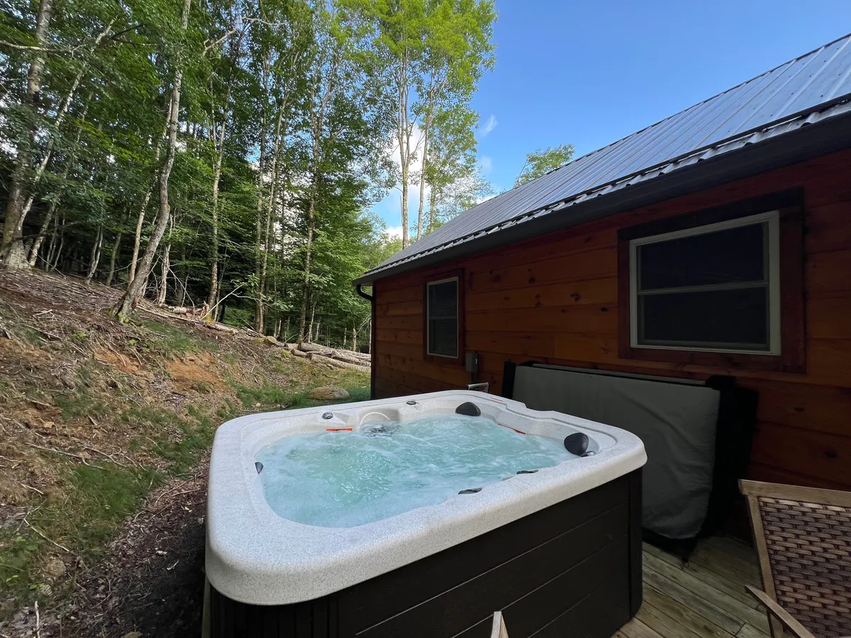 An inviting hot tub rests on a deck surrounded by trees, with a view of the house exterior in the background. The setting is tranquil, ideal for unwinding after a day of outdoor activity, with the promise of sunset views and the sounds of nature.