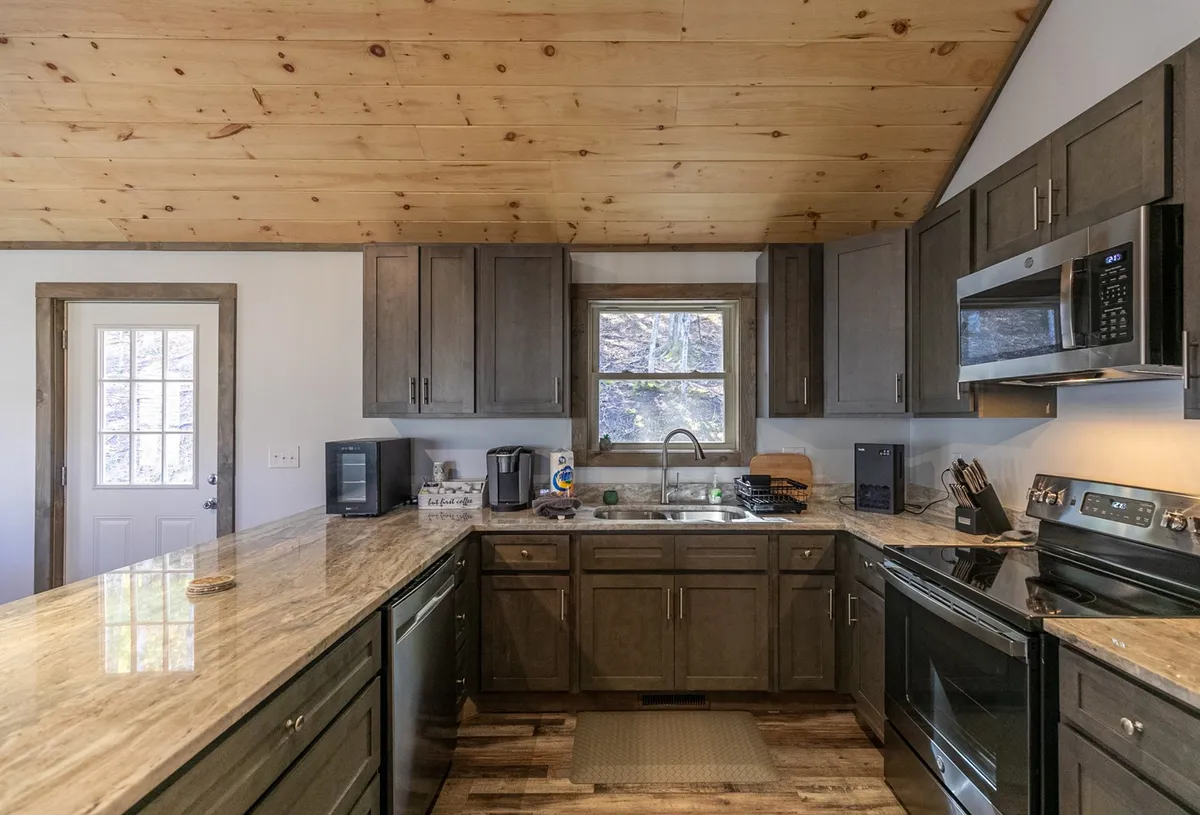 A cozy kitchen scene featuring a large central island with a polished countertop, modern stainless steel appliances, and cabinetry with a rustic wood finish. Large windows invite natural light, and the open design connects the kitchen to a broader living area, emphasizing togetherness and functionality.
