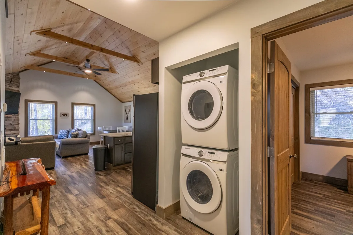 The image features a spacious interior with a washer and dryer, wooden beams, and a stone fireplace, all within an open layout. The setting is warm and inviting, suggesting both modern convenience and rustic charm.