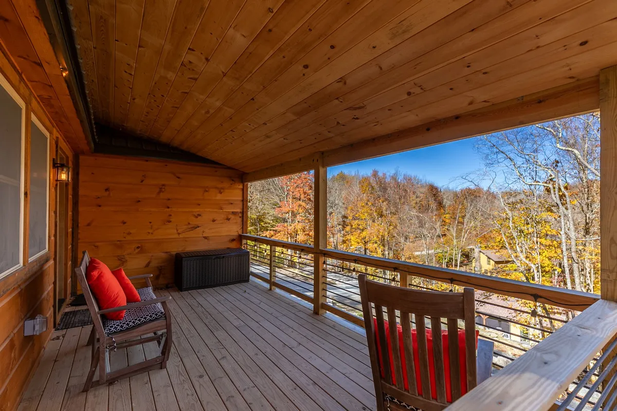 A spacious wooden deck adorned with vibrant red cushions offers a comfortable seating area overlooking a backdrop of brilliant autumn trees. The deck is positioned to capture expansive views and invites guests to relax, sip coffee, or enjoy the outdoors.