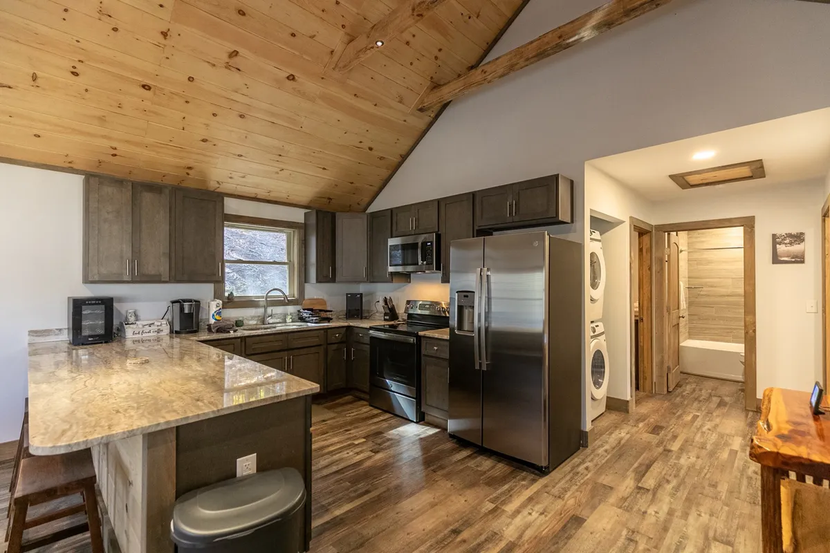 A spacious, inviting kitchen with a gleaming granite countertop, stainless steel appliances, and richly-toned wooden cabinetry. The open layout connects seamlessly to an adjacent living space, suggesting an environment perfect for social cooking and entertaining. High-quality kitchen essentials are visible, underscoring the well-equipped nature of the space.