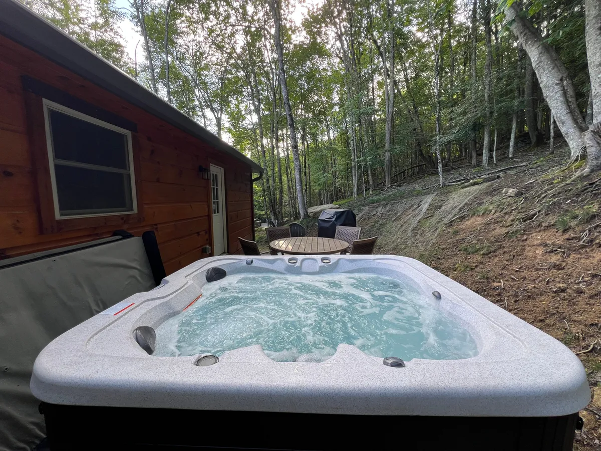 A tranquil setting featuring a bubbling hot tub positioned next to a rustic wooden cabin, surrounded by lush trees. The outdoor seating area invites guests to relax, dine, or socialize, while the natural backdrop highlights the immersive mountain atmosphere. The image is equally evocative of both daytime coffee or evening stargazing experiences.