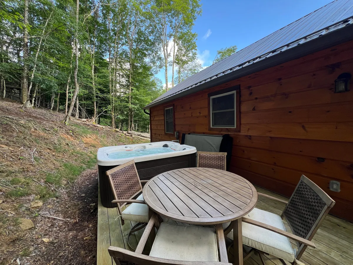 This inviting scene features a hot tub nestled amidst trees, a wooden outdoor table perfect for morning coffee or evening meals, and hints of a fire area for cozy gatherings. The setting feels private and harmonious with the surrounding forest, capturing the essence of both adventure and relaxation.
