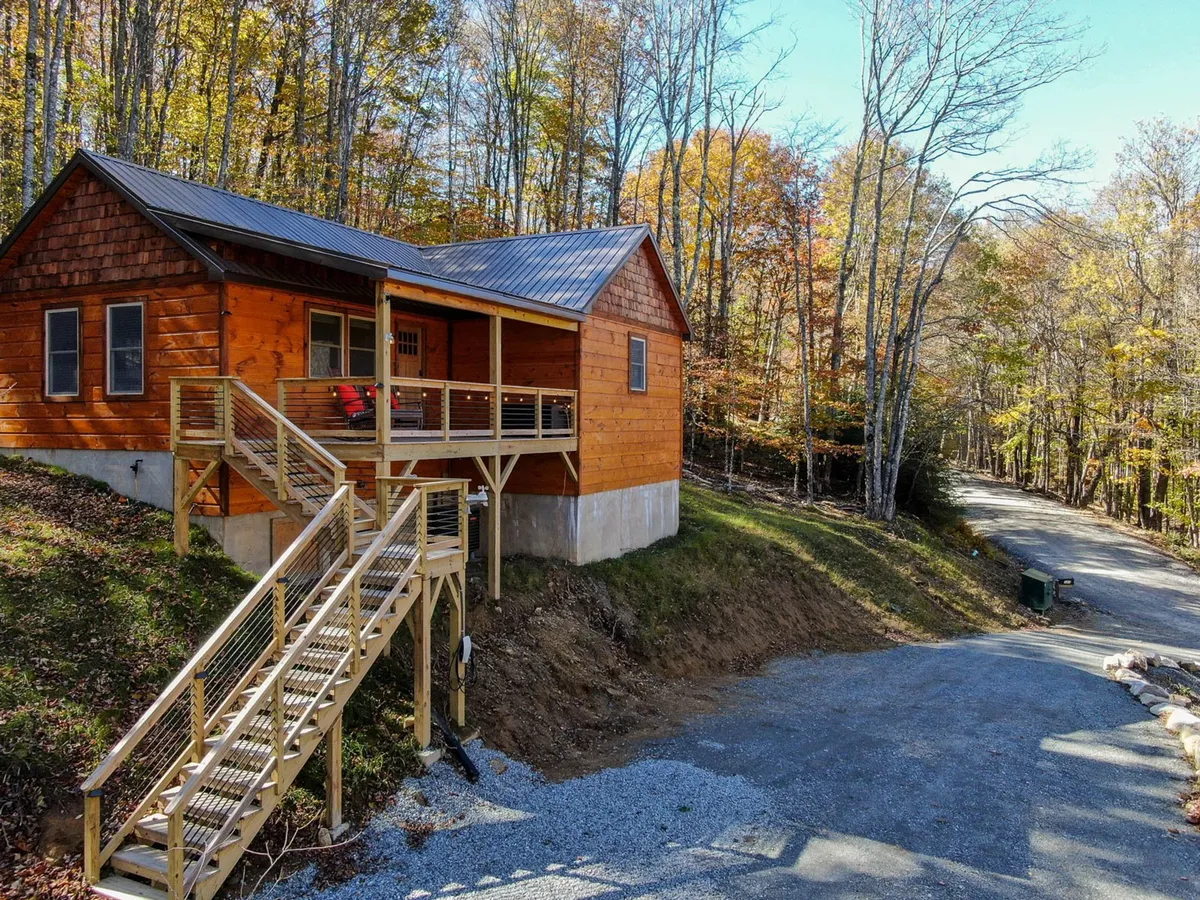 The exterior of Whiskey Rock features a charming wooden façade, a staircase leading up to the porch, and a brilliant display of colorful autumn foliage in the background. The scene suggests a welcoming arrival and connection to nature's beauty.
