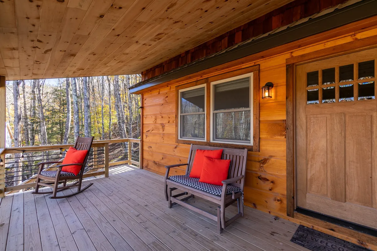 A pair of classic wooden rocking chairs with bright orange cushions sit on a rustic porch, framed by a wooden door and immersed in the surrounding natural landscape. The setting invites quiet moments and social conversation against a mountain backdrop.