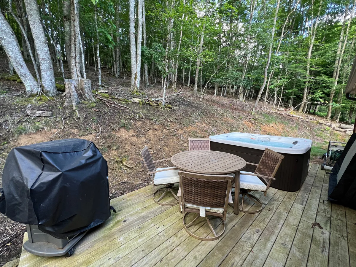 This image captures a spacious wooden deck featuring an inviting hot tub, an outdoor dining set, and a gas grill. The deck is surrounded by natural beauty, with serene mountain views and plenty of space for relaxation and gatherings. The ambiance suggests both daytime and evening enjoyment, from barbecuing to stargazing.